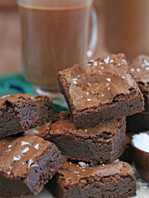 A pile of Salted Fudge Brownies cut into squares next to a bowl of salt a glass of hot chocolate.