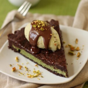 A slice of Pistachio Marzipan Brownie Tart on a white plate covered with Pistachio Ice Cream and Hot Fudge.