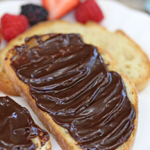 Slice of toast covered in chocolate spread with plain bread and fruit in the background.