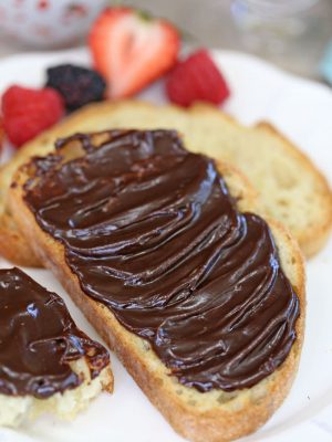 Slice of toast covered in chocolate spread with plain bread and fruit in the background.