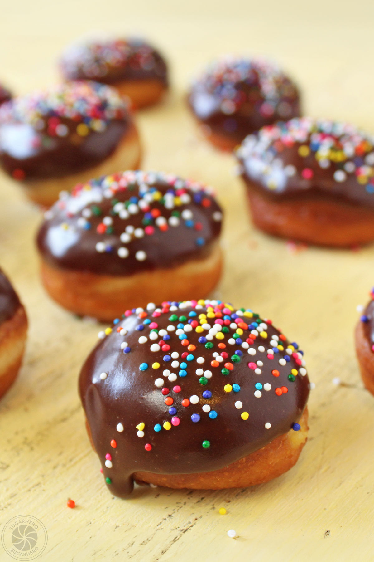 Several Easter Egg Doughnuts drying on wooden surface with multi-colored sprinkles on top.