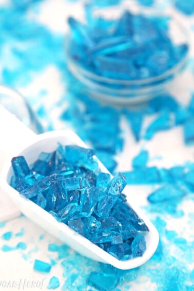 White scoop filled with shards of blue rock candy, on a white surface with candy scattered around.