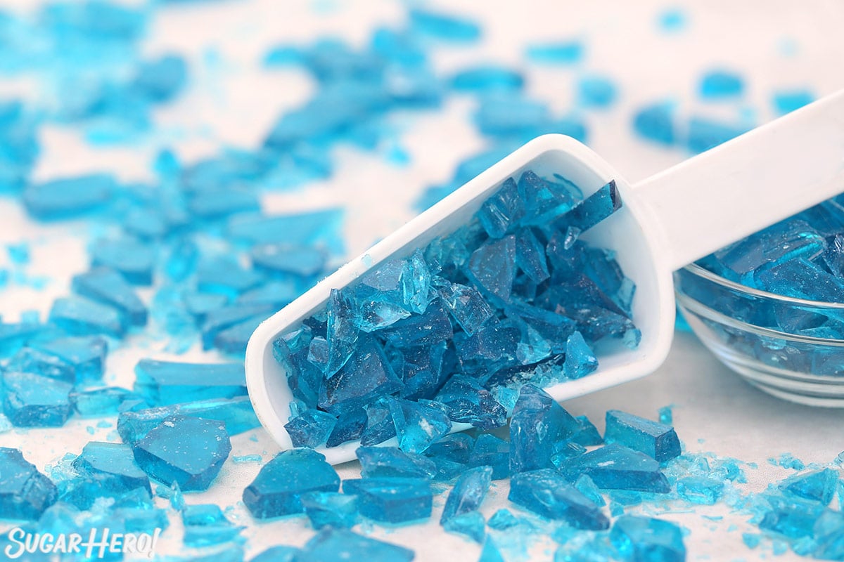 White scoop filled with shards of blue rock candy on a white surface.