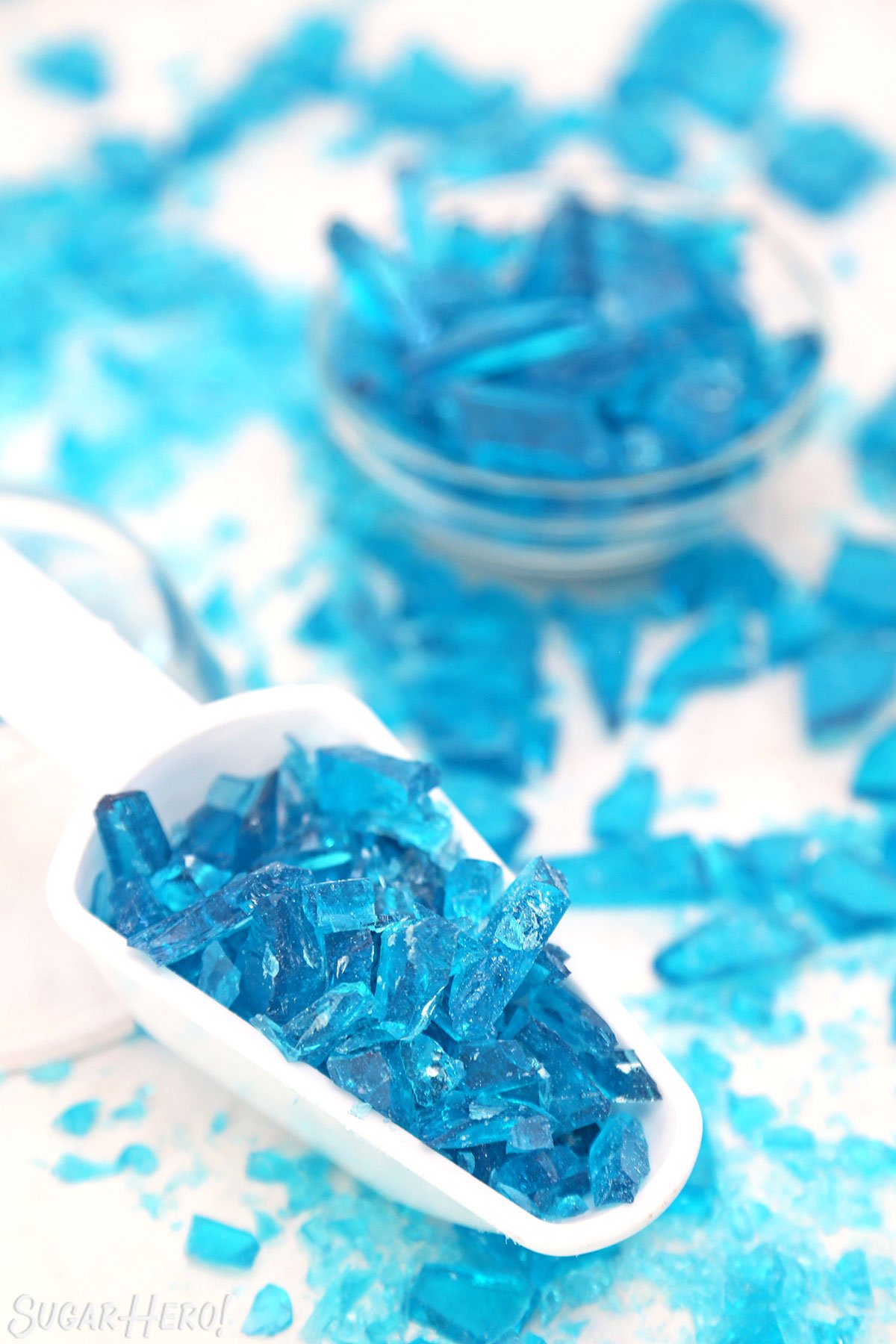 White scoop filled with shards of blue rock candy, on a white surface with candy scattered around.