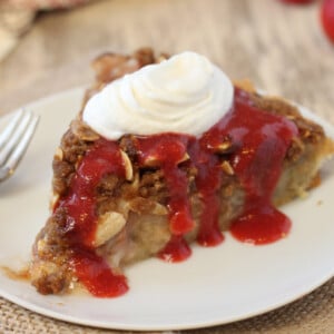 A slice of Rhubarb Streusel Tart on a white plate with strawberry sauce and whipped cream.