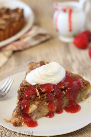 A slice of Rhubarb Streusel Tart on a white plate with strawberry sauce and whipped cream.