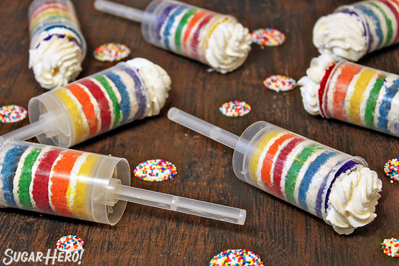 Rainbow Cake Push-Up Pops on their sides on a wooden surface with rainbow-topped candies scattered around.