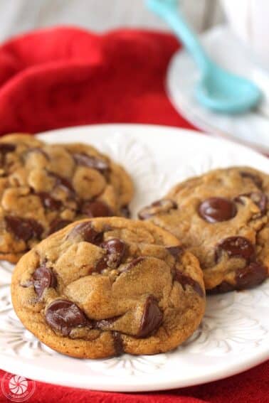 Three gingerbread chocolate chip cookies on a round white plate with a red napkin and blue spoon in the background.