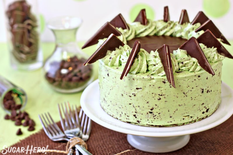 Mint chocolate chip cake on white cake stand, with forks next to it and jars of chocolate chips in the background