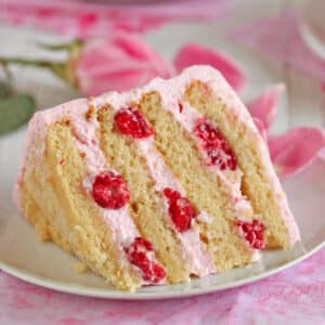 Slice of Raspberry-Rose Cake on a white plate next to floral napkins, a pink rose and a white fork.