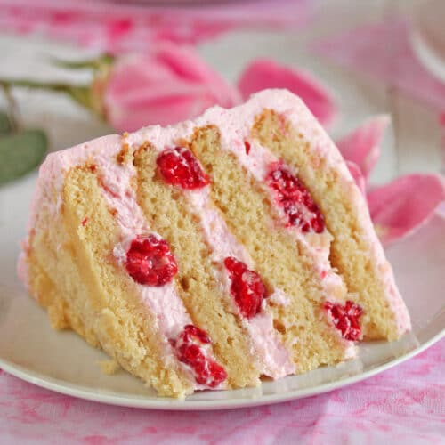 Slice of Raspberry-Rose Cake on a white plate next to floral napkins, a pink rose and a white fork.