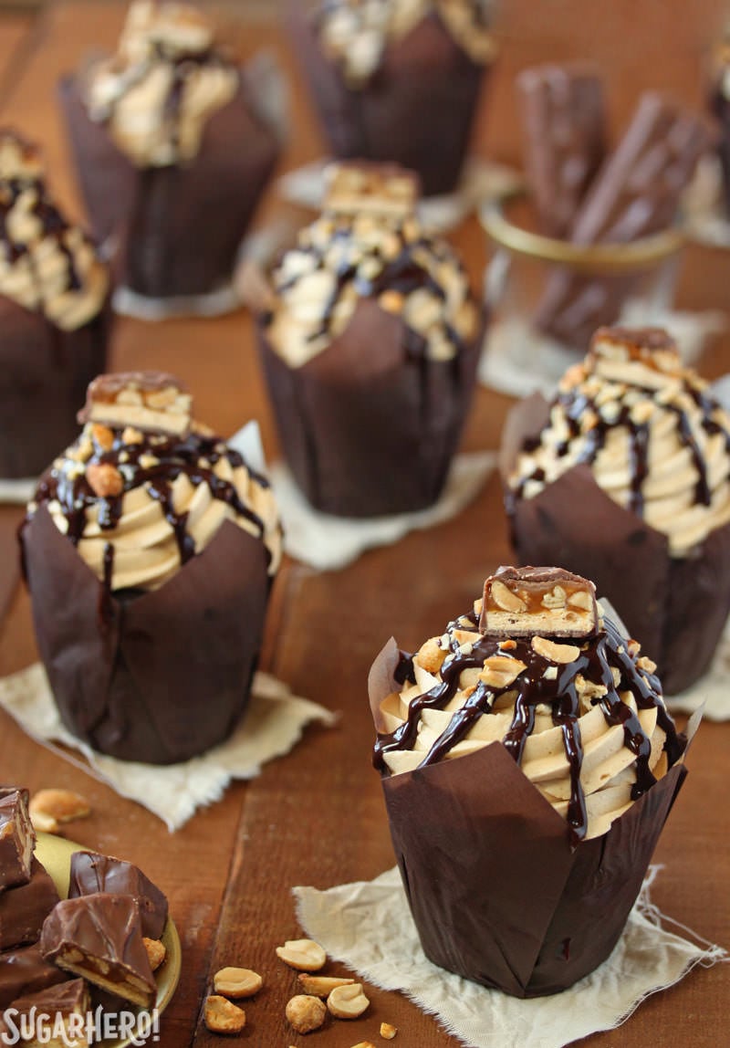 Snickers Cupcakes - In these Snickers Cupcakes, plain chocolate cupcakes are transformed into decadent, candy-bar inspired creations, featuring a peanut caramel filling, fluffy peanut butter frosting, and LOTS of chocolate and candy bars. | From SugarHero.com Snickers Cupcakes - Multiple snicker cupcakes displayed with chocolate dripping down. | From SugarHero.com