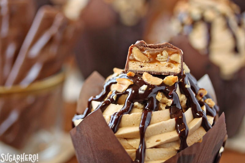 Snickers Cupcakes - In these Snickers Cupcakes, plain chocolate cupcakes are transformed into decadent, candy-bar inspired creations, featuring a peanut caramel filling, fluffy peanut butter frosting, and LOTS of chocolate and candy bars. | From SugarHero.com Snickers Cupcakes - A shot of the top of a cupcake with a piece of snickers as a topper. | From SugarHero.com