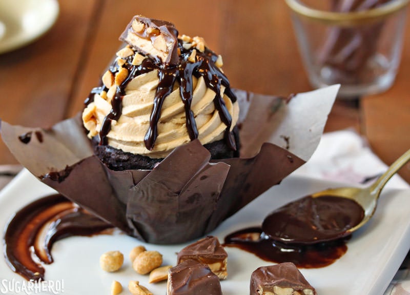Snickers Cupcakes - In these Snickers Cupcakes, plain chocolate cupcakes are transformed into decadent, candy-bar inspired creations, featuring a peanut caramel filling, fluffy peanut butter frosting, and LOTS of chocolate and candy bars. | From SugarHero.com Snickers Cupcakes - A cupcake displayed on a plate with bits of snickers, and a spoon dipped in chocolate. | From SugarHero.com