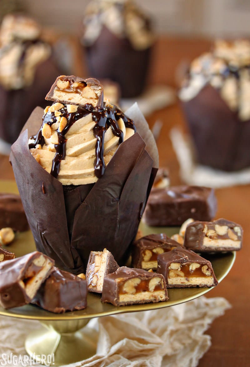 Snickers Cupcakes - In these Snickers Cupcakes, plain chocolate cupcakes are transformed into decadent, candy-bar inspired creations, featuring a peanut caramel filling, fluffy peanut butter frosting, and LOTS of chocolate and candy bars. | From SugarHero.com Snickers Cupcakes - A cupcake displayed on a cake stand with bits of snickers around it. | From SugarHero.com