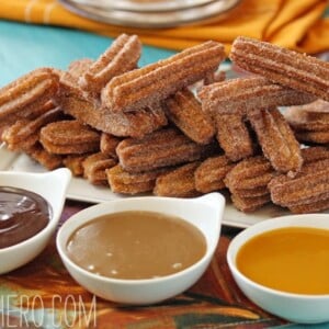 Plate stacked with homemade churros next to 3 small bowls of chocolate, dulce de leche and mango sauces.