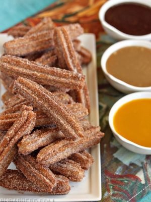 Stacked plate of homemade churros next to 3 small bowls of chocolate, dulce de leche and mango sauces.
