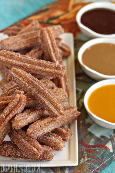 Stacked plate of homemade churros next to 3 small bowls of chocolate, dulce de leche and mango sauces.