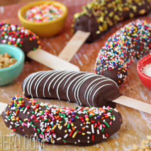 6 chocolate-dipped frozen bananas and an assortment of toppings to roll them in on a wooden surface.