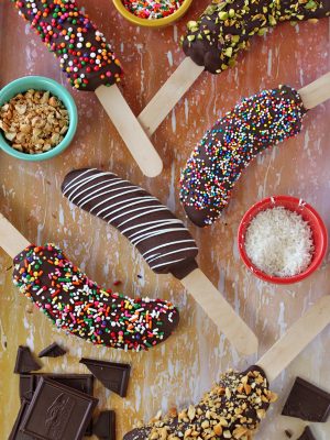 Overhead shot of 6 chocolate-dipped frozen bananas and an assortment of toppings to roll them in on a wooden surface.