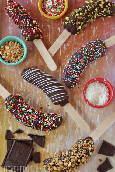 Overhead shot of 6 chocolate-dipped frozen bananas and an assortment of toppings to roll them in on a wooden surface.
