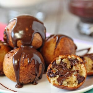 A Close up of a pile of Deep Fried Chocolate Chip Cookie Dough balls on a white plate drizzled with chocolate and cut open to show interior.