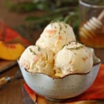 3 large scoops of Peach Honey Ice Cream in a stoneware bowl with honey and peaches in the background.