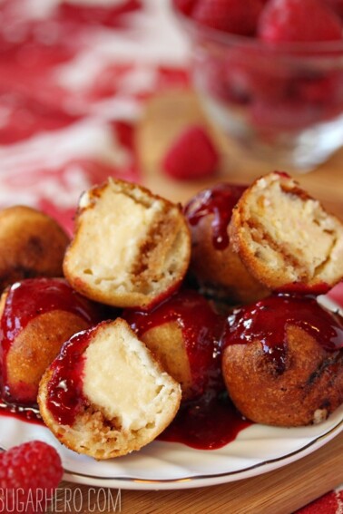 7 deep-fried cheesecake bites piled on a white plate, drizzled with raspberry sauce and cut to show center filling.