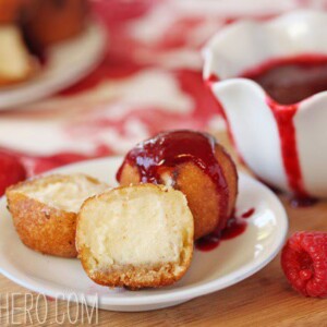 1deep-fried cheesecake bite cut to show center and another drizzled with raspberry sauce on a white plate with sauce in the background.