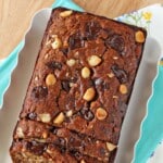 A loaf of Banana Coconut Macadamia Nut Bread on a white tray and teal napkin with a few cut slices in the front.