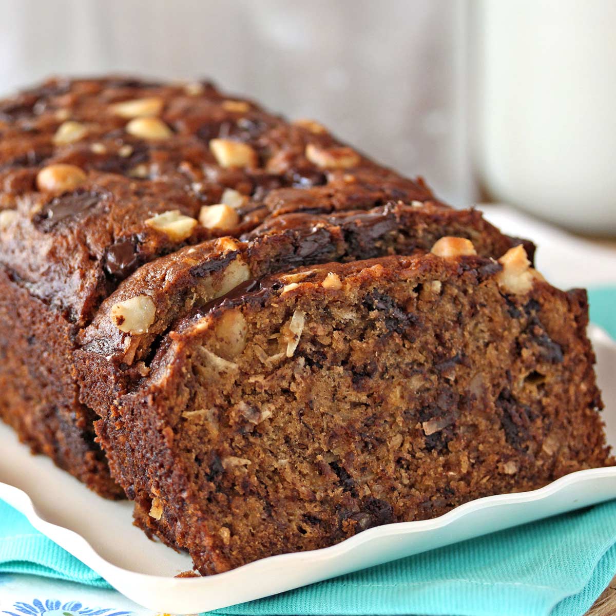 A loaf of Banana Coconut Macadamia Nut Bread on a white tray and teal napkin with a few cut slices in the front.