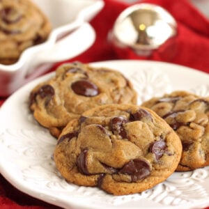 Three gingerbread chocolate chip cookies on a white round plate with a few more cookies in a scalloped square serving dish on a red napkin in the background.