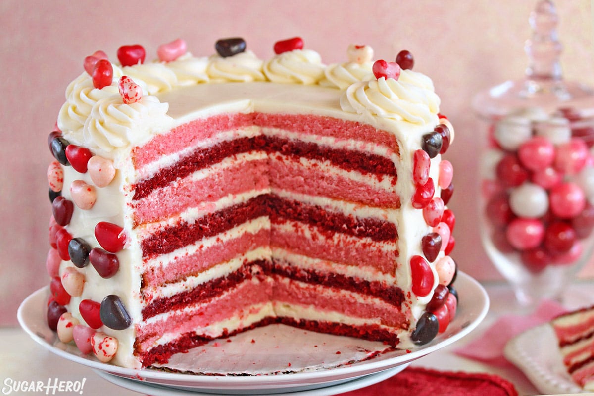 Layer cake with several slices taken out, showing pink and red cake layers with cream cheese frosting.