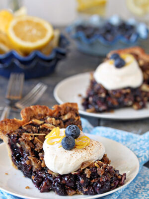 Two slices of blueberry crumble pie topped with whipped cream, fresh blueberries, and lemon zest.