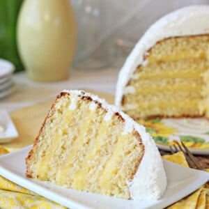 A slice of Lemon Coconut Snowball Cake on a small square plate with the rest of the cake in the background.
