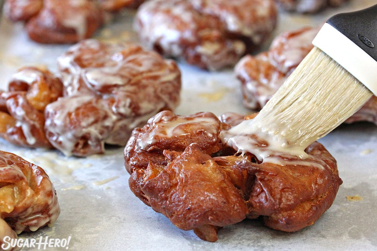 Apple Cider Fritters | From SugarHero.com Apple Cider Fritters being glazed on parchment paper.