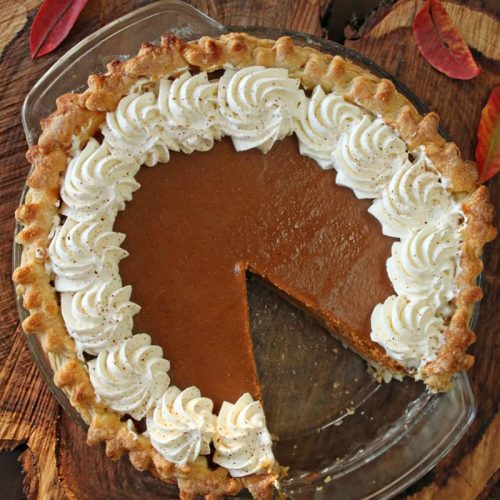 Dulce de Leche Pumpkin Pie with slice missing on a piece of wood with fall leaves.