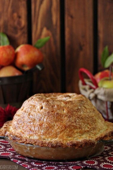 Mile High Apple Pie with crisp golden crust on a red napkin with baskets of apples in the background.