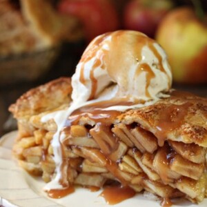 Slice of mile high apple pie on a white plate topped with vanilla ice cream and caramel sauce.