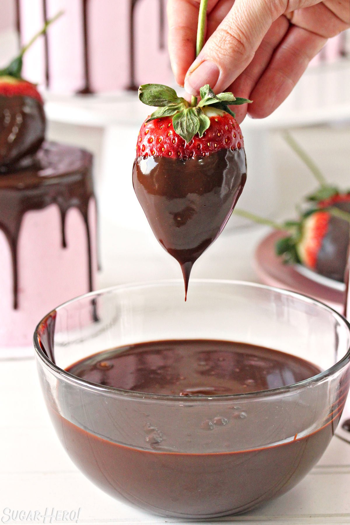 Hand holding a large strawberry by the stem, dripping melted chocolate back into a chocolate bowl below.