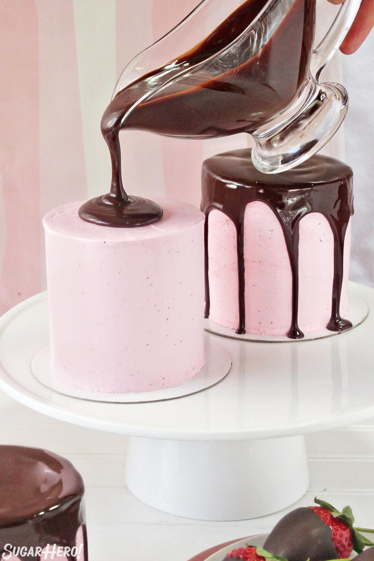 Pouring shiny chocolate ganache on top of a pink-frosted miniature cake.