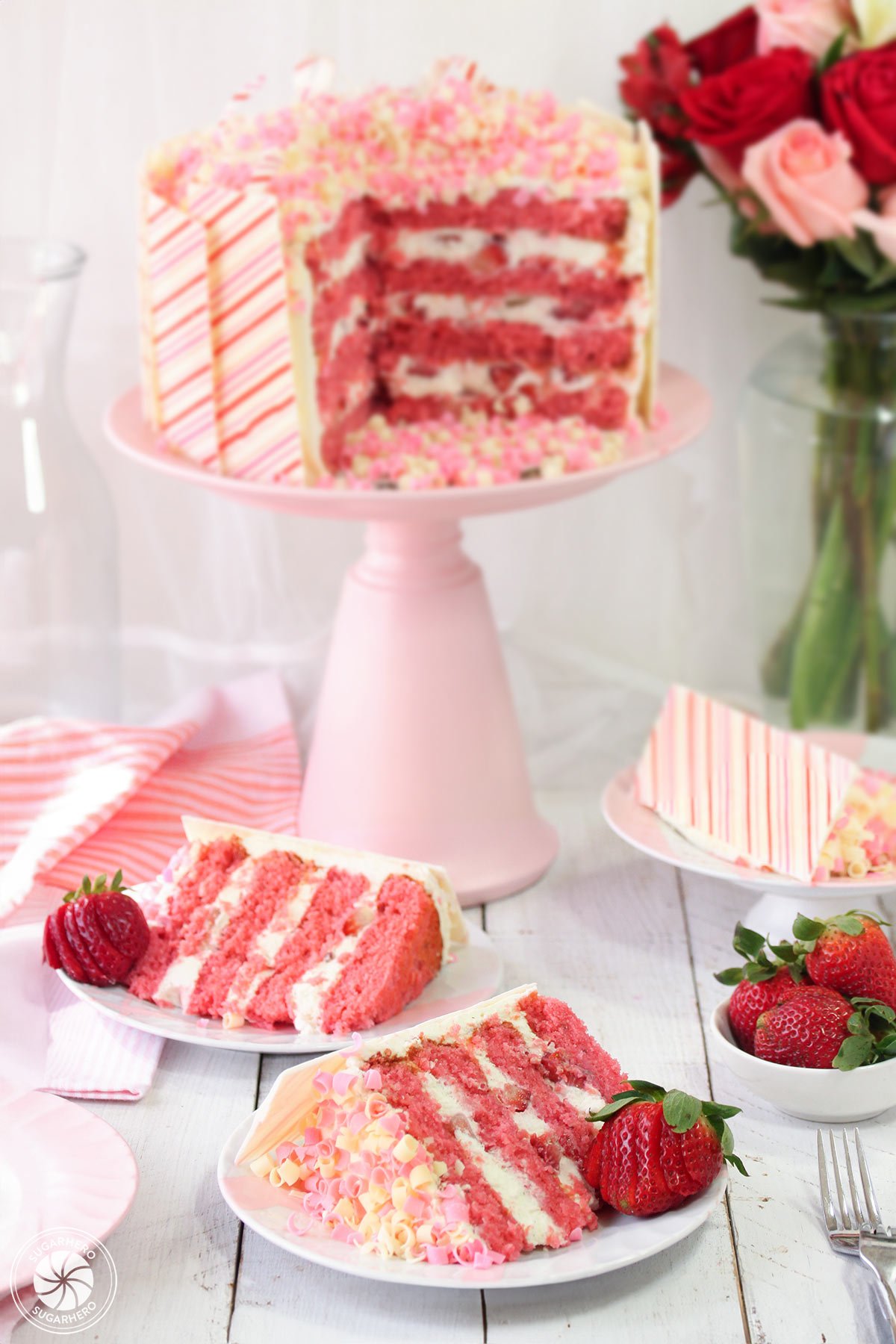 Strawberries and Cream Layer Cake - vertical image of cake on a pink cake stand, with several pieces of cake on plates around the base | From SugarHero.com