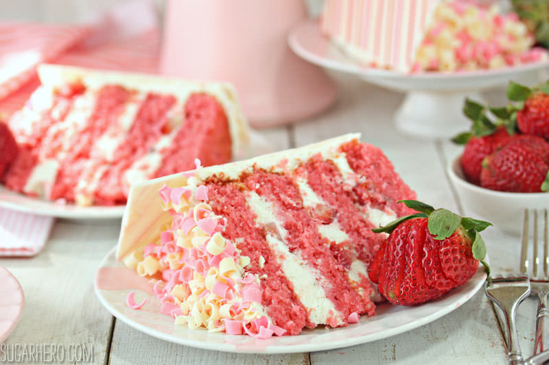 Strawberries and Cream Layer Cake - close-up of a slice of strawberry cake with pink and white chocolate curls on top | From SugarHero.com