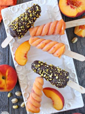 An assortment of Peach Passion Pops on a white rectangular platter next to peaches and pistachios.