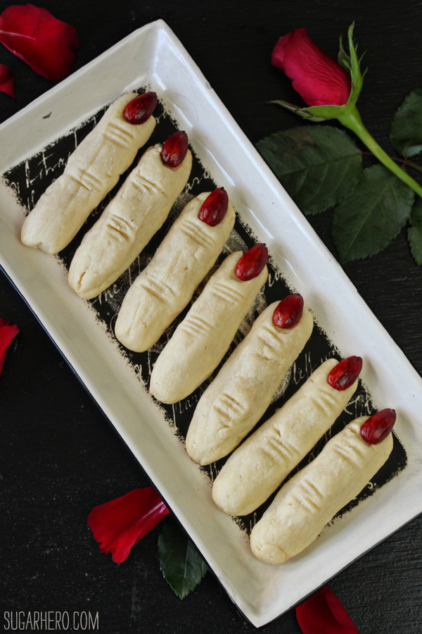 White rectangular plate holding 7 almond witch finger cookies next to a red rose.