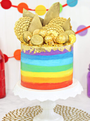 Gold-Topped Rainbow Cake on a white cake stand in front of colorful bunting in the background.