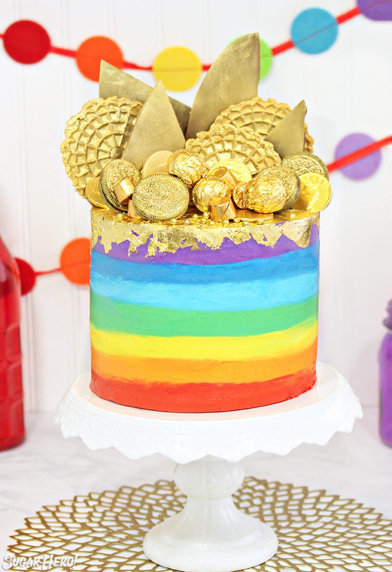 Gold-Topped Rainbow Cake on a white cake stand in front of colorful bunting in the background.