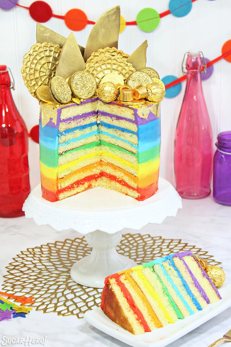 Gold-Topped Rainbow Cake sliced open to reveal the rainbow frosting layers inside, on a white cake stand.