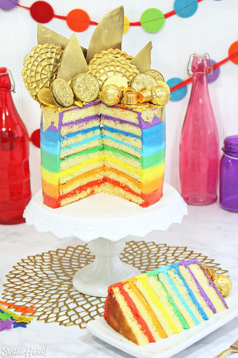 Pot of Gold Rainbow Cake - with EIGHT rainbow layers and sparkling edible gold treats on top! | From SugarHero.com Gold-Topped Rainbow Cake sliced open to reveal the rainbow frosting layers inside, on a white cake stand.