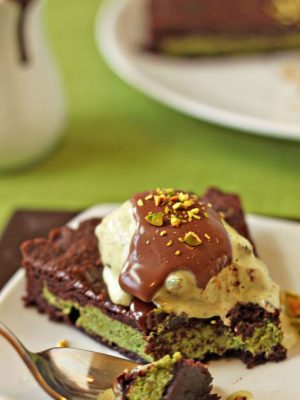 A slice of Pistachio Marzipan Brownie Tart with a bite removed by a fork on a white plate covered with Pistachio Ice Cream and Hot Fudge.